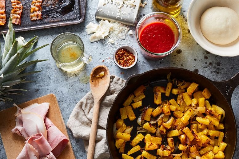 Pineapple in a pan, ham on a wooden cutting board, wooden spoon, and dough in a bowl.